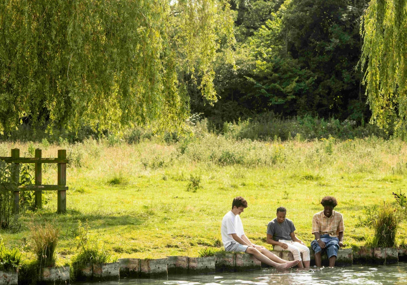 Three students chatting and sitting in a field with their feet in the river.